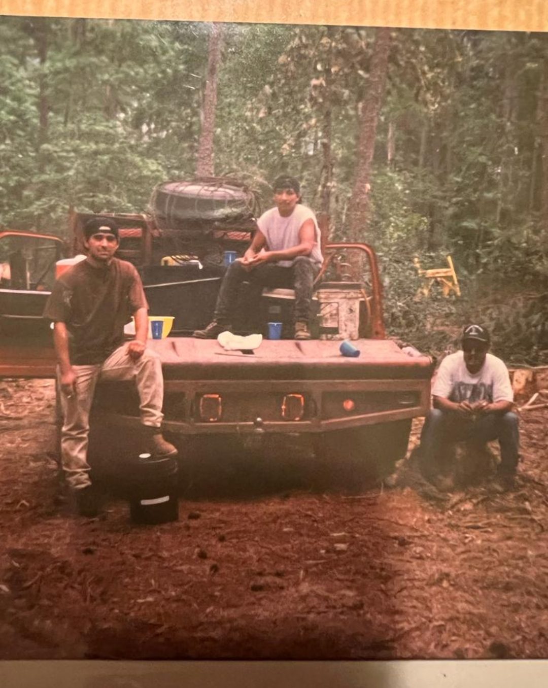 Three men relax on a vehicle in a forested area, surrounded by trees and outdoor gear.