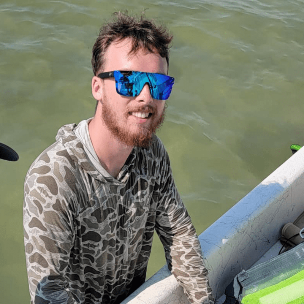 Smiling man in sunglasses and a camo shirt stands by a boat on water, showcasing a relaxed outdoor lifestyle.