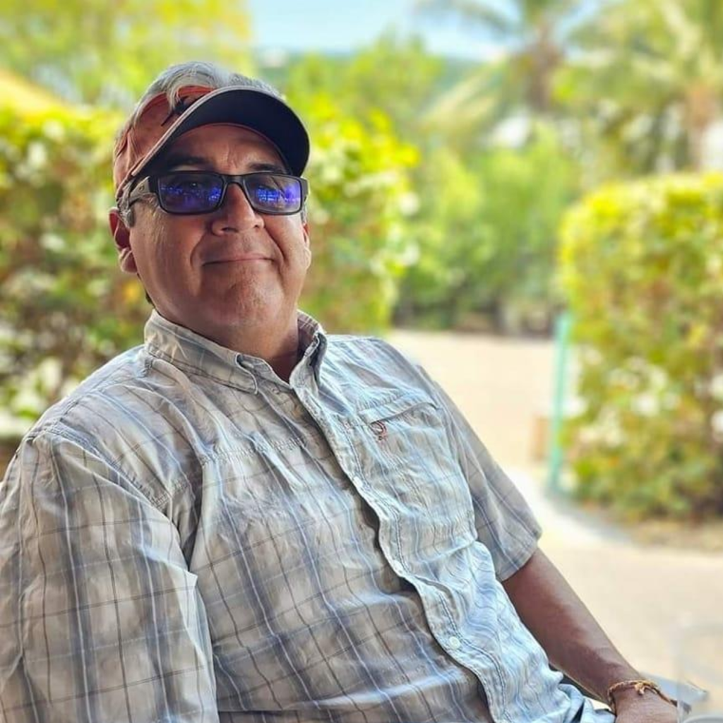 Smiling man in a plaid shirt and sunglasses sits outdoors surrounded by greenery, conveying a relaxed atmosphere.